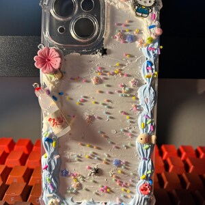 Puede incluir: Funda de tel&eacute;fono transparente decorada con una variedad de adornos coloridos. La funda presenta un borde azul claro con un dise&ntilde;o de remolino y est&aacute; adornada con una flor rosa, una botella en miniatura y un dije de Hello Kitty.