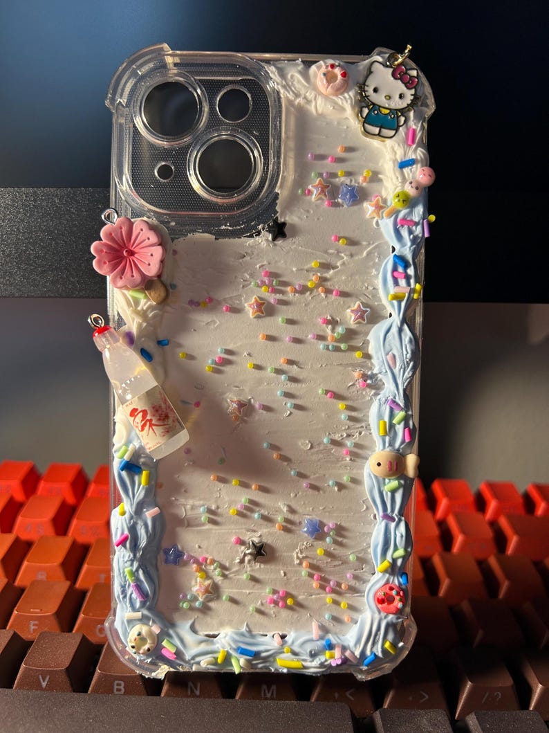 Puede incluir: Funda de tel&eacute;fono transparente decorada con una variedad de adornos coloridos. La funda presenta un borde azul claro con un dise&ntilde;o de remolino y est&aacute; adornada con una flor rosa, una botella en miniatura y un dije de Hello Kitty.