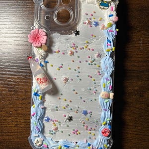 Puede incluir: Funda de tel&eacute;fono transparente decorada con glaseado blanco, detalles de glaseado azul y chispas de colores. Adornada con dijes que incluyen una flor rosa, una botella en miniatura, un dije de Hello Kitty y un pez peque&ntilde;o.