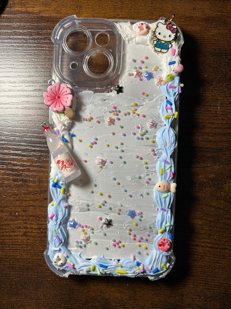 Puede incluir: Funda de tel&eacute;fono transparente decorada con glaseado blanco, detalles de glaseado azul y chispas de colores. Adornada con dijes que incluyen una flor rosa, una botella en miniatura, un dije de Hello Kitty y un pez peque&ntilde;o.