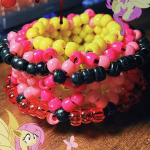 May include: A colorful beaded cuff bracelet featuring yellow, pink, red, and black beads. The bracelet is a stackable design, with multiple rows of beads. The image also includes cartoon graphics of a pony, butterflies, and apples.