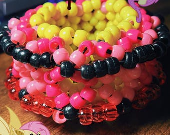 May include: A colorful beaded cuff bracelet featuring yellow, pink, red, and black beads. The bracelet is a stackable design, with multiple rows of beads. The image also includes cartoon graphics of a pony, butterflies, and apples.