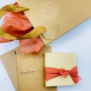 May include: A brown paper gift bag with a white tag that says "To" and "From:"  The bag is tied with a gold ribbon and has a white circle with the letter "K" inside. The bag is sitting on a brown envelope with the words "Message for you" written on it. There is a small, square, brown box with a red ribbon tied around it.