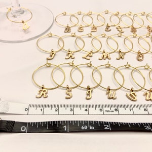 May include: A set of 18 gold-colored wine glass charms with letter charms. The charms are arranged in alphabetical order from A to U. The charms are on a white surface with a ruler for scale.