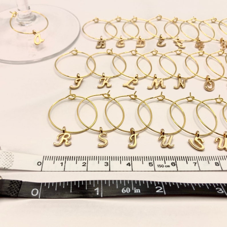 May include: A set of 18 gold-colored wine glass charms with letter charms. The charms are arranged in alphabetical order from A to U. The charms are on a white surface with a ruler for scale.