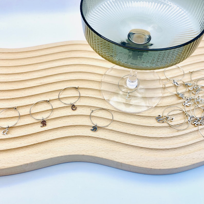 May include: A set of silver wine glass charms with different letter charms. The charms are on a wooden surface with a glass goblet in the background.