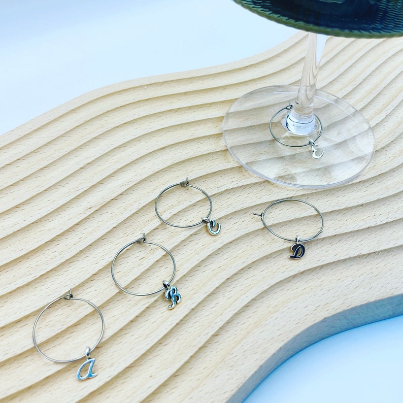 May include: Four silver wine glass charms with letter charms hanging from each. The letters are A, B, C, and D. The charms are on a wooden surface.