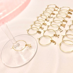 May include: A set of 20 gold-colored metal wine glass charms with letter charms. The charms are arranged in a triangle shape on a white surface. One charm is on a wine glass.
