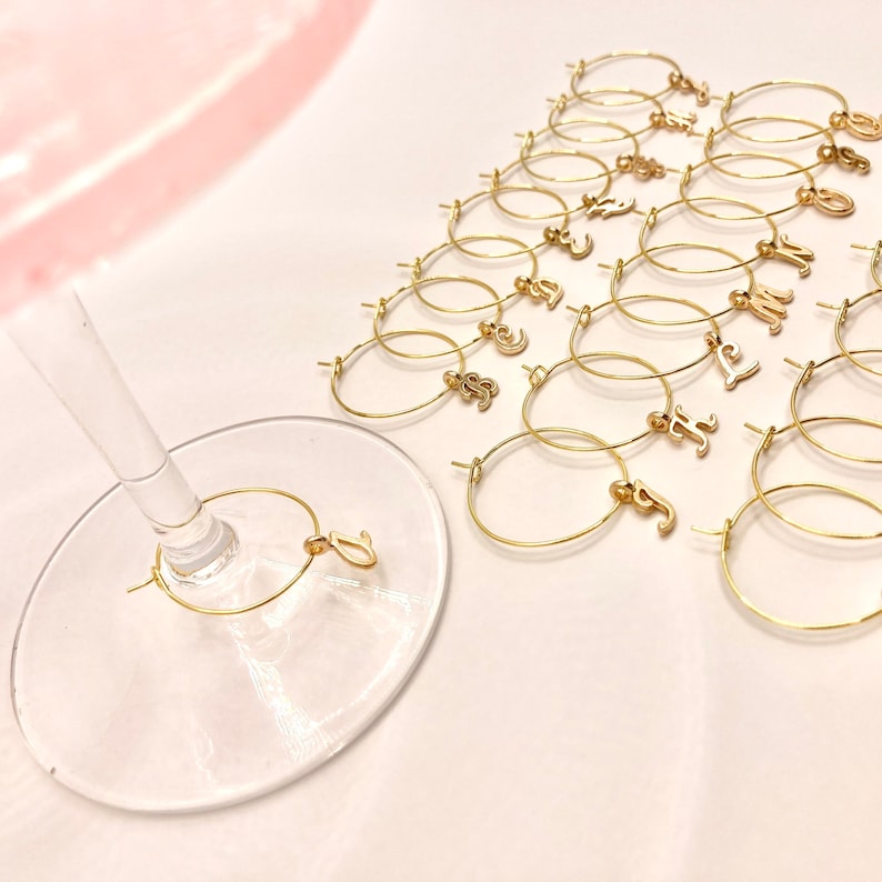 May include: A set of 20 gold-colored metal wine glass charms with letter charms. The charms are arranged in a triangle shape on a white surface. One charm is on a wine glass.
