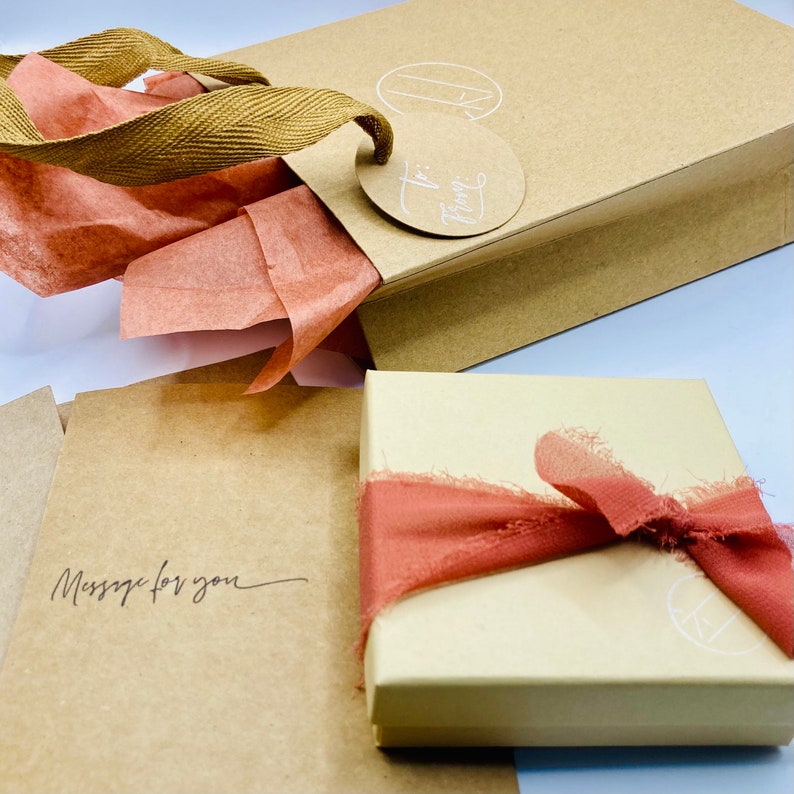 K&ouml;nnte beinhalten: Eine braune Papiert&uuml;te mit einem braunen Band und einem runden Etikett mit der Aufschrift "To From". Die T&uuml;te ist mit rosa Seidenpapier gef&uuml;llt. Eine kleine braune Schachtel mit einem wei&szlig;en Kreis und einem rosa Band liegt auf einer braunen Oberfl&auml;che. Die Schachtel tr&auml;gt eine handschriftliche Nachricht mit der Aufschrift "Message for you".