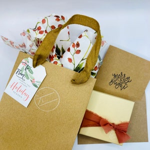 May include: A brown paper gift bag with a white and red floral design tissue paper inside. The bag has a brown ribbon handle and a white tag that says "From:" and "Wishing you a happy Holiday Season". The bag is sitting on a brown envelope with a white card that says "Merry & Bright".