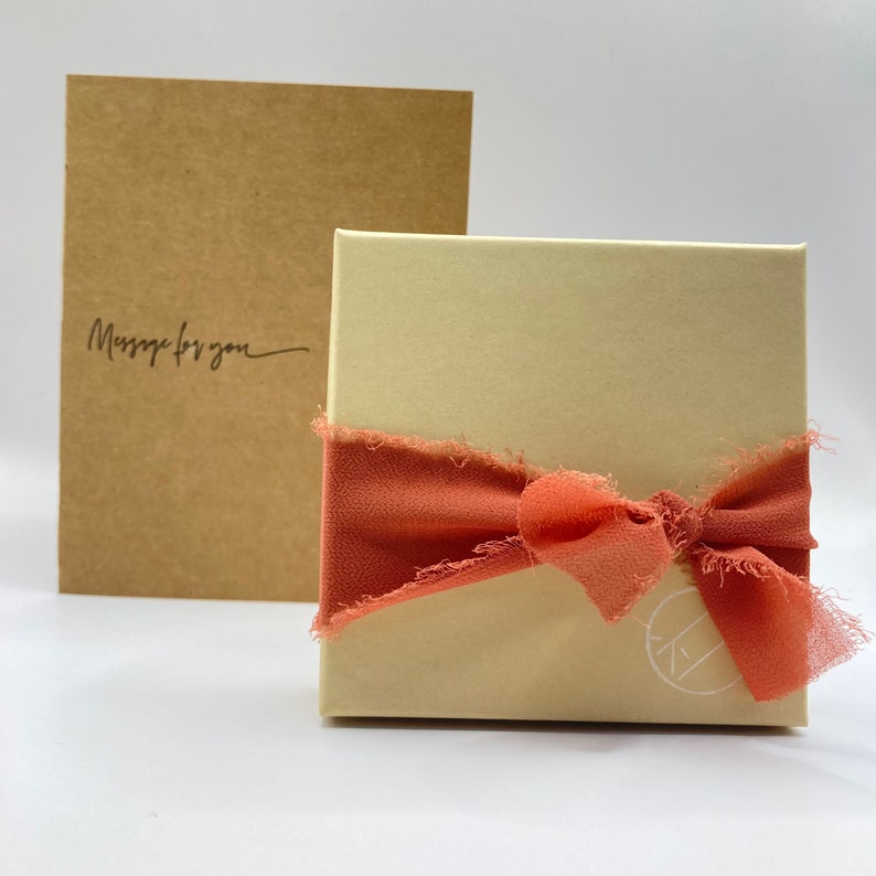 May include: A cream-colored gift box with a coral-colored ribbon tied in a bow. The box is sitting on a white surface. The text "Message for you" is written on a brown card behind the box.
