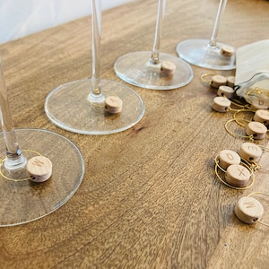 Könnte beinhalten: Ein Satz Holz-Wein-Glas-Anhänger mit goldenen Ringen und gravierten Buchstaben auf jedem Anhänger. Die Anhänger befinden sich in einem kleinen weißen Beutel mit Kordelzug.
