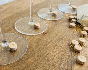 May include: A set of wooden wine glass charms with gold rings and letters engraved on each charm. The charms are in a small white drawstring bag.