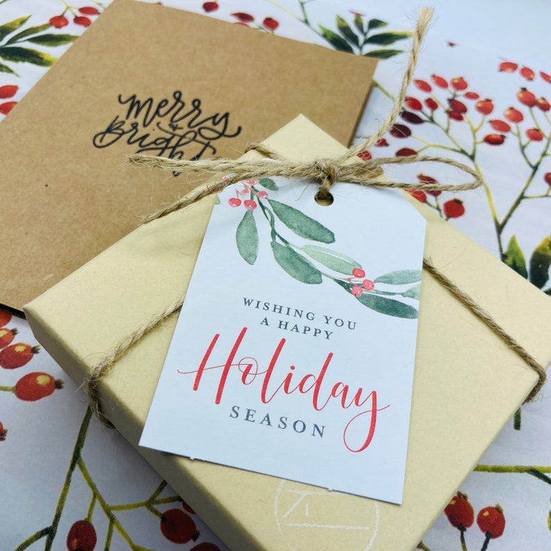 May include: A gift box wrapped in brown paper with a white gift tag tied with twine. The gift tag has a watercolor illustration of green leaves and red berries with the text "Wishing you a happy Holiday Season".