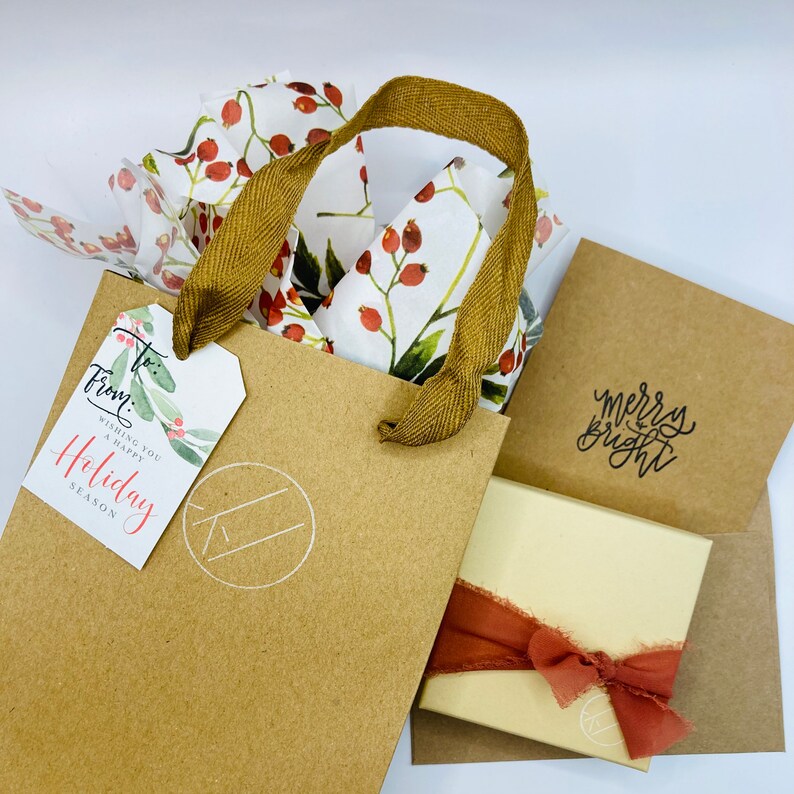 K&ouml;nnte beinhalten: Eine braune Papiert&uuml;te mit wei&szlig;em und rotem Blumenmuster-Seidenpapier im Inneren. Die Tasche hat einen braunen Bandgriff und ein wei&szlig;es Etikett mit der Aufschrift "Von:" und "Wir w&uuml;nschen Ihnen eine frohe Weihnachtszeit". Die Tasche liegt auf einem braunen Umschlag mit einer wei&szlig;en Karte, auf der "Frohe und helle Weihnachten" steht.