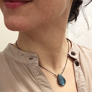 Könnte beinhalten: Halskette mit einem tropfenförmigen, blauen Labradorit-Anhänger. Der Edelstein hängt an einer schwarzen Kordel mit kleinen Silberperlen. Die Halskette wird über einem hellrosa Hemd getragen.