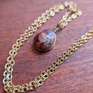 Könnte beinhalten: Eine Vintage-Halskette in Goldoptik mit einem Anhänger. Der Anhänger ist ein ovaler Stein mit roten, braunen und grauen Farben. Die Kette hat ein doppeltes Glieddesign. Die Halskette liegt auf einer Holzoberfläche.