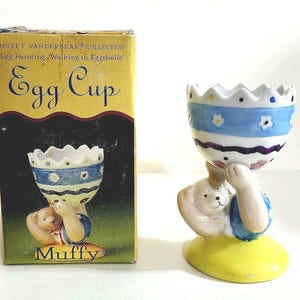 Può includere: Un portauovo in ceramica della collezione Muffy Vanderbear. Il portauovo è tenuto da una statuina di orso e presenta un design blu e bianco. La scatola recita "Egg Cup" e "Egg Painting, Walking in Eggshells."