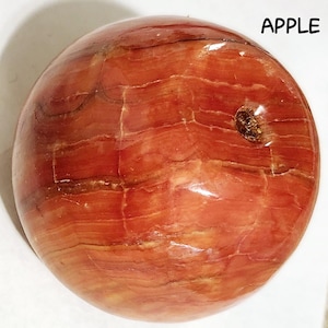 May include: A polished, spherical stone with a reddish-brown color and a layered, natural pattern. The word "APPLE" is printed in black, uppercase letters on the upper right side of the image.