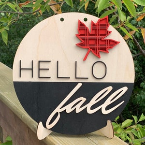 Peut inclure: Une pancarte ronde en bois avec les mots "HELLO fall" en lettres noires. La pancarte est peinte en noir et blanc et a une feuille d'érable rouge à carreaux attachée au sommet.