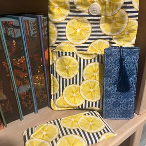 May include: A fabric sleeve with a yellow lemon and navy blue stripe pattern, featuring a white button closure and a front pocket. Beside it is a rectangular blue fabric case with a dark blue tassel, and a matching lemon-patterned fabric pouch.