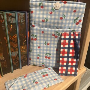 May include: A fabric book sleeve with a light blue and white gingham pattern featuring red cherry prints, secured by a white button. Included is a matching rectangular fabric bookmark with a blue tassel and a separate small rectangular fabric pouch.