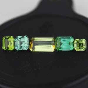 Może przedstawiać: Kolekcja pięciu kamieni szlachetnych w różnych odcieniach zieleni i żółci. Kamienie są prostokątne i kwadratowe, o różnych rozmiarach. Kamienie szlachetne są prezentowane na czarnej powierzchni.