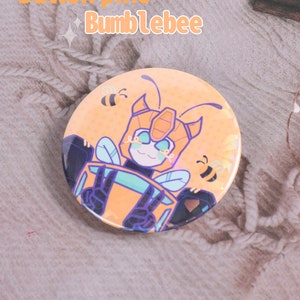 Peut inclure: Un badge bouton de 58 mm avec un personnage de dessin animé jaune et noir de Bumblebee Transformer faisant un signe de paix. Le badge a une finition brillante et holographique.