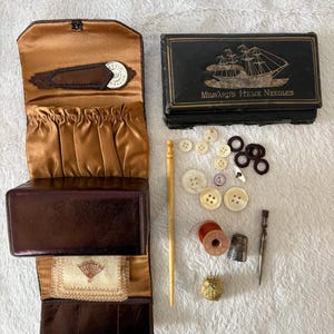 Puede incluir: Un kit de costura de cuero marrón con interior de satén dorado, una caja negra con la etiqueta Milward's Helix Needles y un barco, botones blancos y oscuros, una aguja de madera, un carrete de hilo rojo, un dedal y una herramienta de costura.