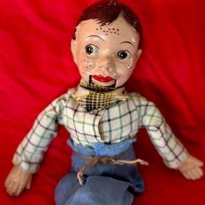 Puede incluir: Un muñeco de ventrílocuo con cara pintada, pelo castaño y pecas. El muñeco lleva una camisa blanca de manga larga con un patrón de cuadros verdes y negros, un pañuelo de cuello a cuadros amarillos y negros y pantalones azules atados.