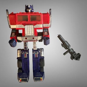 Puede incluir: Una figura de acción de robot en colores rojo, azul y plateado con un torso de cabina de camión, cabeza azul y extremidades articuladas. La figura presenta una parrilla y parachoques plateados, junto a un rifle de plástico gris.