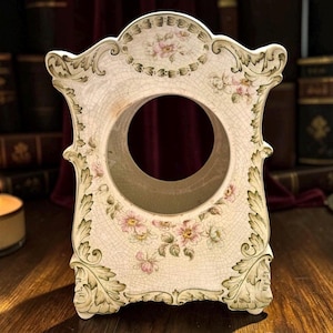 May include: An ornate ceramic clock housing featuring a cream-colored base with a visible crackle glaze finish. The structure is decorated with painted green acanthus leaf scrollwork and delicate pink floral clusters surrounding a central circular opening.