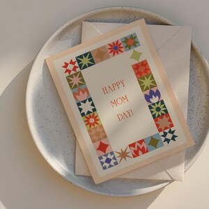 May include: A greeting card with the text "HAPPY MOM DAY!" surrounded by a colorful floral border. The card is on top of a cream-colored envelope, resting on a speckled ceramic plate.