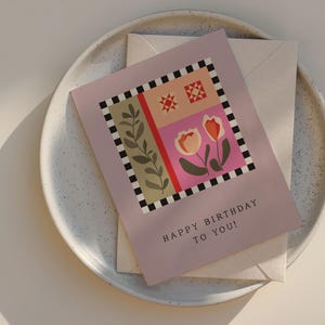 May include: A birthday card with a floral design and the text "HAPPY BIRTHDAY TO YOU!" is placed on a speckled ceramic plate with a matching envelope. The card features a pink background with a colorful geometric pattern.