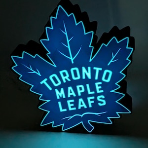 Puede incluir: Un logotipo azul brillante de los Toronto Maple Leafs con forma de hoja de arce. El nombre del equipo se muestra en blanco, centrado en la hoja. Los bordes de la hoja están delineados con una luz azul brillante.