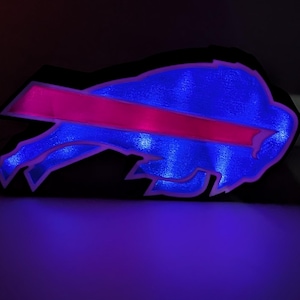 Op de afbeelding: Een gloeiend blauw en roze Buffalo Bills logo, verlicht tegen een donkere achtergrond. Het logo toont een blauwe buffelkop met een roze streep erover. De randen van het logo zijn omlijnd met helder blauw licht.