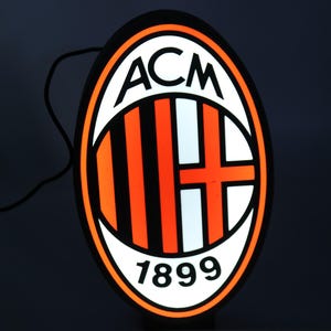 May include: Illuminated sign featuring the ACM Milan logo. The oval-shaped sign has a black border with the team's emblem in the center. The emblem includes the letters "ACM", vertical orange and black stripes, a white cross, and the year "1899".