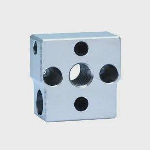 Puede incluir: Un bloque metálico plateado, rectangular, con múltiples orificios circulares de varios tamaños. El bloque tiene un orificio central roscado y varios orificios más pequeños en sus lados y parte superior, lo que sugiere un componente mecánico.