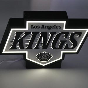 May include: A black and white illuminated Los Angeles Kings logo sign. The sign features the team name and a crown emblem. The sign is lit with white LED lights, and has a power cord.