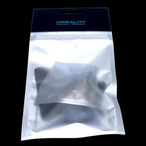 May include: A transparent plastic bag with a black header bearing the text "CREALITY". Inside the bag is a dark object, likely a 3D printing component. The bag is sealed and has a hanging hole.
