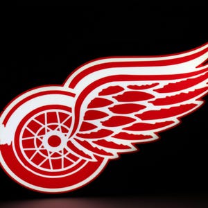 May include: Red and white Detroit Red Wings logo on a black background. The logo features a stylized red wing with white accents and a red wheel with white spokes. The design is a symbol of the Detroit Red Wings hockey team.