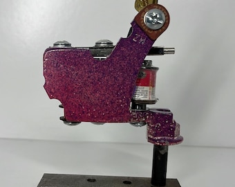 May include: A purple and silver tattoo machine with a rectangular base. The machine has a coil and needle assembly, with a brass screw and a leather-like accent. The machine is designed for professional tattoo artists.