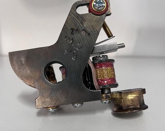 May include: A silver tattoo machine with a red and gold coil. The machine has a brass base and a silver needle bar. The machine is marked with the text "ZM 220".