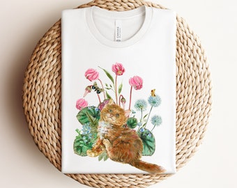 May include: A white t-shirt with a watercolor illustration of a ginger cat sitting in a bed of pink and white flowers. The cat is looking up with a playful expression. The flowers are surrounded by green leaves and there are two small bees flying around the flowers.