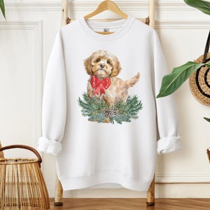 May include: White sweatshirt with a watercolor illustration of a small, light brown dog wearing a red bow and standing in a wreath of green pine branches with pine cones.