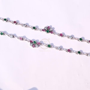Op de afbeelding: Twee zilveren enkelbandjes met een bloemmotief. Elk enkelbandje heeft afwisselend roze, groene en heldere edelstenen. De enkelbandjes worden gepresenteerd op een wit oppervlak. Het bloemmotief bevindt zich in het midden.