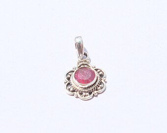 May include: A silver pendant featuring a round, red gemstone. The gemstone is set within a decorative, flower-shaped frame with detailed patterns. The pendant includes a small loop at the top for a chain, typical of jewellery.