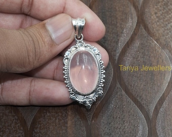 May include: A silver pendant with a delicate, ornate design features a light pink oval-shaped rose quartz gemstone. The pendant is held in a hand against a brown wooden background.