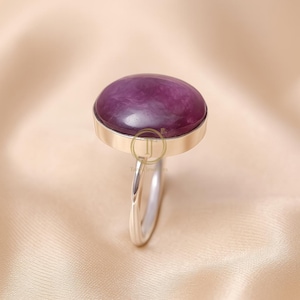 May include: A silver ring featuring a large, oval-shaped, deep purple gemstone. The gemstone is set in a silver bezel, and the band is a simple, polished silver. The ring is displayed against a soft, neutral background.